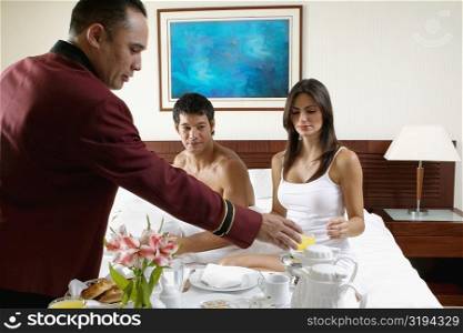 Side profile of a waiter serving a young woman and a mid adult man breakfast in bed