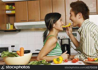 Side profile of a young man feeding a slice of yellow bell pepper to a young woman at a kitchen counter