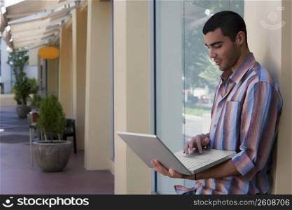 Side profile of a young man leaning against the wall and using a laptop