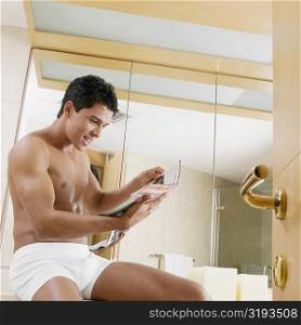 Side profile of a young man sitting in the bathroom and reading a magazine