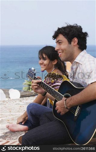 Side profile of a young man sitting with a young woman and playing a guitar