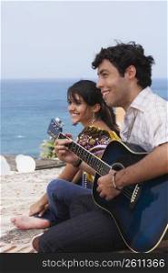 Side profile of a young man sitting with a young woman and playing a guitar