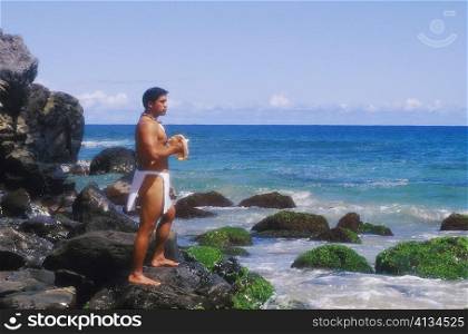 Side profile of a young man standing on a rock and holding a conch shell, Hawaii, USA
