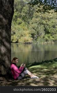 Side profile of a young woman leaning against a tree and text messaging
