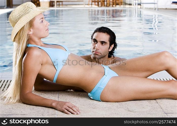 Side profile of a young woman lying down at the poolside with a mid adult man in the swimming pool