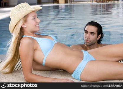 Side profile of a young woman lying down at the poolside with a mid adult man in the swimming pool