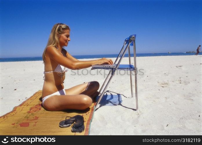 Side profile of a young woman working on a laptop on the beach