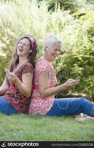 Side profile of two mature women sitting back to back in a park and listening to MP3 players
