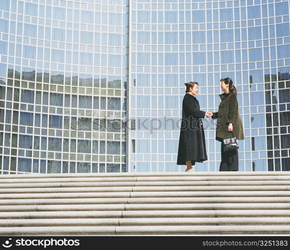 Side profile of two young women standing and shaking hands in front of a building