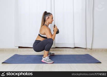Side view full body of fit young female in sportswear squatting with kettlebell while training muscles during fitness workout at home. Athletic woman doing squats with weight