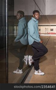 Side view full body of self assured young black male millennial with dark hair in trendy outfit and sneakers leaning on glass wall in passage and looking at camera. Fashionable young African American guy leaning on glass wall in passage
