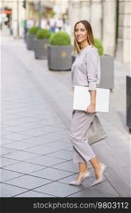 Side view full body of self assured young female entrepreneur with blond hair, in classy clothes and high heels with laptop in hand smiling while walking on paved street in city. Confident well dressed businesswoman with netbook strolling in city