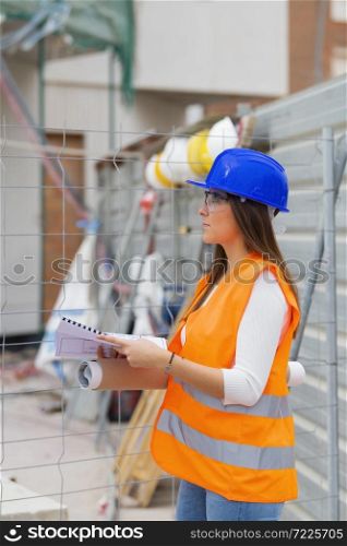 Side view of a beautiful young architecture student wearing safety gear holding blueprints and checking a construction site. Work and apprenticeship concept.. Architecture student holding blueprints checks a construction site