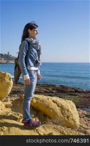 Side view of a daydreaming woman wearing casual clothes standing on seashore while looking away to the horizon in a bright day