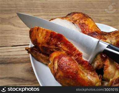 Side view of a freshly oven roasted whole chicken being sliced by large kitchen knife in white serving dish placed on rustic wood