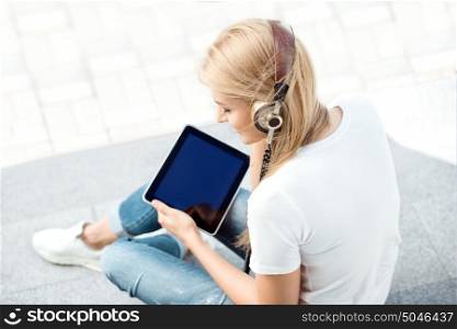 Side view of a happy young woman, listening to the music in vintage music headphones, surfing internet on a tablet pc and sitting on stairs against urban city background.