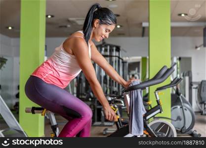 Side view of a latin sportswoman looking down adjusting a stationary bike while doing exercise at gym.. Side view of a young latin sportswoman adjusting a stationary bike while doing exercise at gym