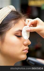 Side view of a makeup artist covers female eye with a cotton pad using airbrush making makeup foundation on her face in a beauty salon. Side view of a makeup artist covers female eye with a cotton pad using airbrush making makeup foundation on her face in a beauty salon.
