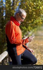 Side view of a senior runner man leaning on fence while testing exercise in a mobile phone outdoors in a sunny day