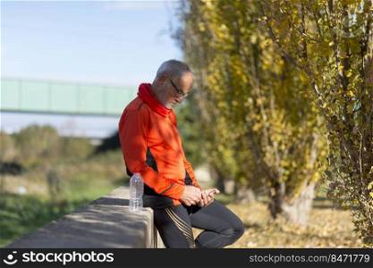 Side view of a senior runner man leaning on fence while testing exercise in a mobile phone outdoors in a sunny day