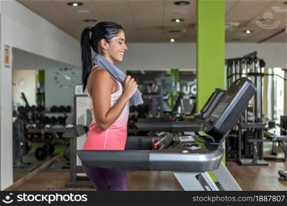 Side view of a young adult woman wearing sportswear walking on a treadmill with a towel around her neck looking to the panel of machine.. Young adult woman wearing sportswear walking on a treadmill with a towel around her neck looking to the panel of machine