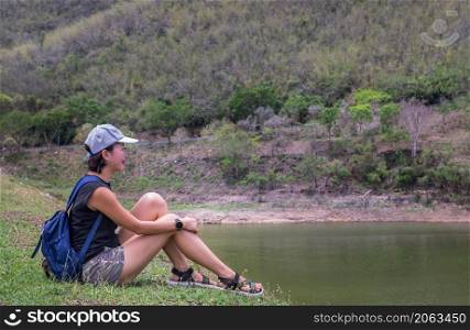 Side view of a young asia woman sitting and enjoying peaceful moment of beautiful view at lake shore with mountains range in background. Holiday travel concept, Rest and enjoyment, lifestyle, Selective focus.