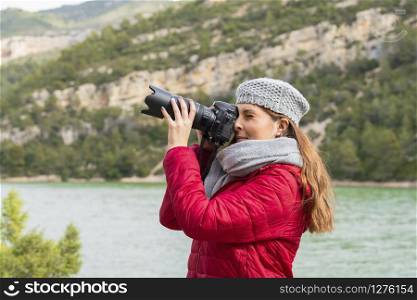Side view of a young beautiful woman wearing a scarf and beret holding a camera and taking a photo of the scenery on an out of focus background. Vacation and hobby concept.. Young photographer taking a photo of the scenery