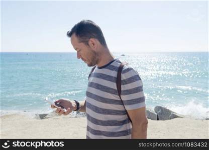 Side view of a young man in t-shirt while using mobile phone on seashore in sunny day