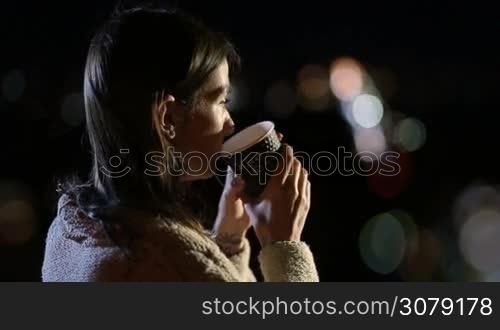 Side view of adorable young long brown hair lady enjoying night city lights while drinking hot fresh coffee from takeawy cup. Beautiful woman with paper cup of hot drink relaxing at summer night outdoors over defocused streetlights background.