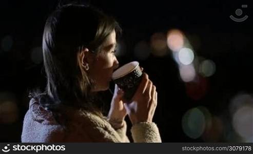 Side view of adorable young long brown hair lady enjoying night city lights while drinking hot fresh coffee from takeawy cup. Beautiful woman with paper cup of hot drink relaxing at summer night outdoors over defocused streetlights background.