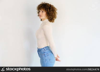 Side view of adult female in beige turtleneck and jeans standing with arms behind back against white background looking away. Emotionless woman standing with hands clasped behind back