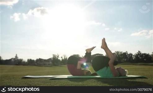 Side view of adult fitness women training outdoors, doing rolling like a ball exercise. Attractive fit ladies practicing yoga and strengthening the abdominals while working out on green grass over beautiful landscape background. Dolly shot.