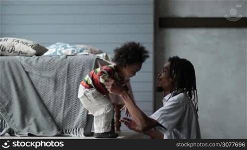 Side view of african american dad with dreadlocks lifting his little mixed race toddler son up in the air over his head in modern apartment. Smiling yong father playing with his cute curly giggling child at home. Slow motion.