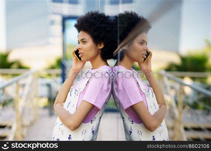 Side view of African American female having phone conversation while standing near glass building on street of city against blurred background. Black woman talking on smartphone near building
