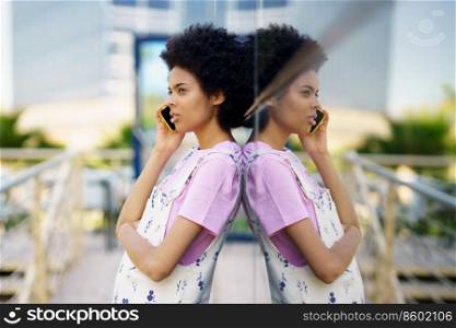 Side view of African American female having phone conversation while standing near glass building on street of city against blurred background. Black woman talking on smartphone near building