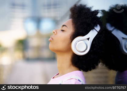 Side view of African American female listening to song in wireless headphones near glass building on street against blurred background. Black woman listening to music on street