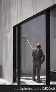 Side view of Asian builder worker using long handle roller brush to applying primer white paint on cement wall in door frame on concrete wall inside of house construction site area