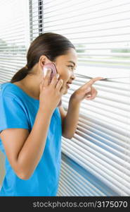 Side view of Asian preteen girl looking through blinds out window while talking on cell phone.
