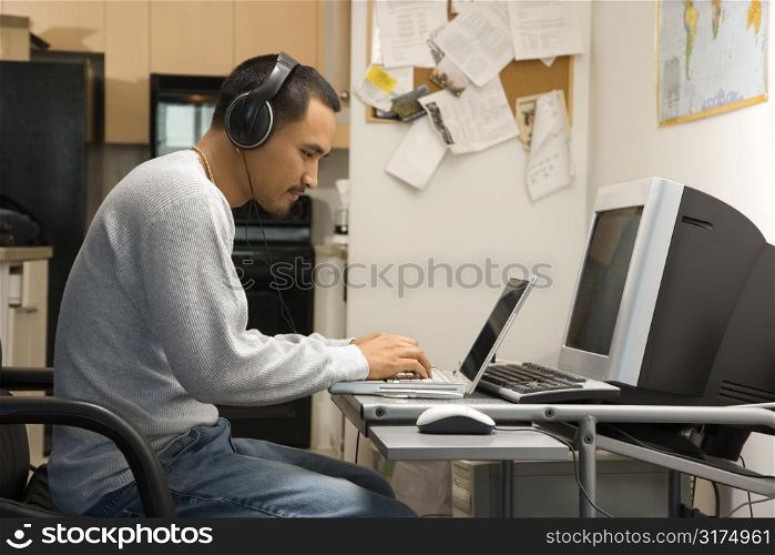 Side view of Asian young adult man sitting at desk typing on laptop wearing headphones.