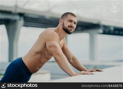 Side view of athletic motivated purposeful bearded sportive bare chested man performs push ups exercise, makes efforts leans on bridge hence looks into distance, has muscular body, works out outside.