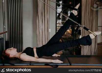 Side view of athletic slender woman doing legs stretching exercises on cadillac reformer in lying position showing body strength and flexibility during pilates training in studio. Sport and fitness. Flexible redhead woman doing legs stretching exercises on cadillac reformer