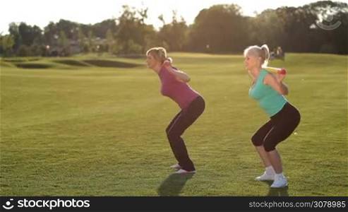 Side view of attractive adult blonde females doing squats workout with body bars on their shoulders on park lawn in glow of amazing sunset. Positive sporty fit senior women in great shape exercising with weights doing squatting outdoors.