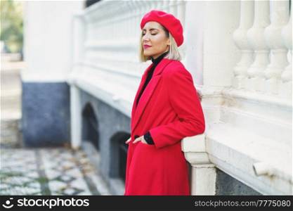 Side view of attractive female in red outfit with beret standing with hands in pockets near white fence on street. Fashionable woman near white fence in urban background