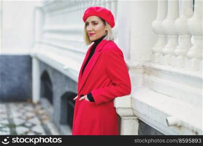 Side view of attractive female with blond hair in read coat and beret standing with hands in pockets near white fence on street. Trendy woman in beret near fence outdoors