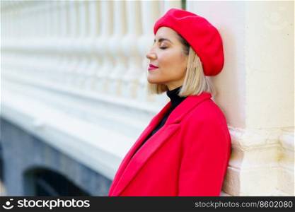 Side view of attractive female with blond hair in red beret leaning on white column with closed eyes near fence while standing on street. Stylish woman in red wear in city