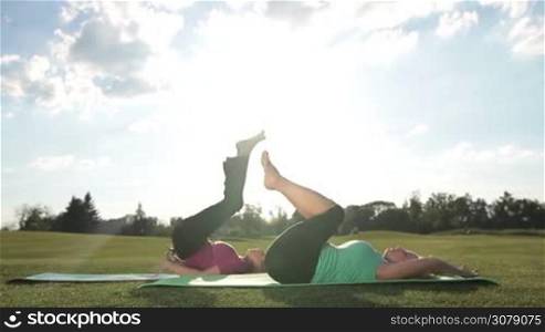 Side view of attractive senior fitness female practicing yoga, stretching in shoulder stand exercise while working out on green grass in rays of light. Dolly shot. Active adult women doing upside down seal pose in park while sitting on exercise mats