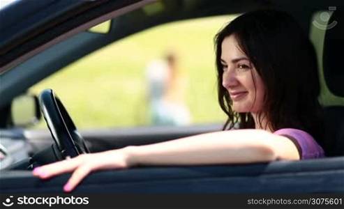 Side view of attractive woman driver sitting in driver&acute;s seat with open window, on the background beautiful long-haired blonde is standing in flower&acute;s field and waving hello