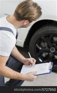 Side view of automobile mechanic writing on clipboard while examining car&rsquo;s wheel in workshop