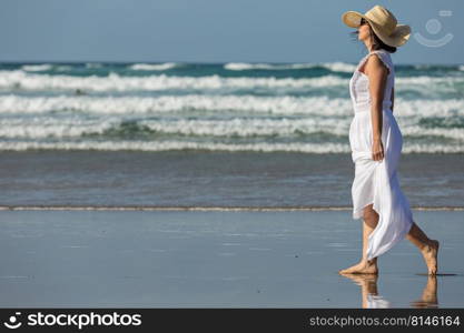 Side view of barefoot female in white maxi dress and straw hat walking on wet sand against sea waves and blue sky while spending weekend day on beach. Woman walking against waving sea