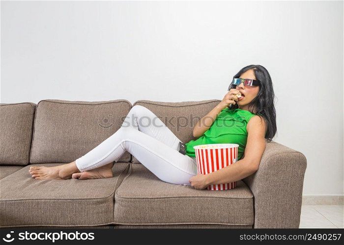 Side view of barefoot Hispanic female in casual clothes and 3D glasses eating fresh popcorn and watching interesting movie while chilling on couch in light living room. Relaxed woman with popcorn watching movie on sofa